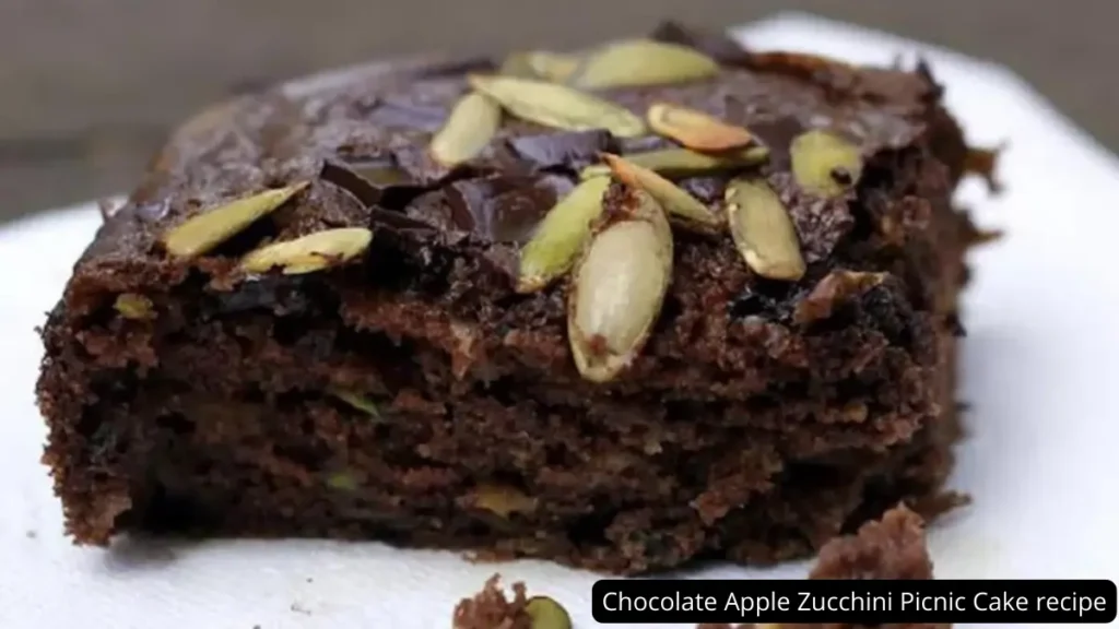 A close-up of a slice of Chocolate Apple Zucchini Picnic Cake topped with dark chocolate chunks and green pepitas, emphasizing its moist texture and rich, dark color.