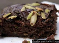 A close-up of a slice of Chocolate Apple Zucchini Picnic Cake topped with dark chocolate chunks and green pepitas, emphasizing its moist texture and rich, dark color.