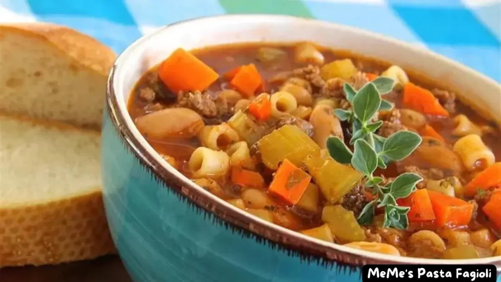 A close-up view of a bowl of Meme's Pasta Fagioli, featuring ditalini pasta, cannellini beans, ground beef, and diced vegetables, with slices of bread in the background.