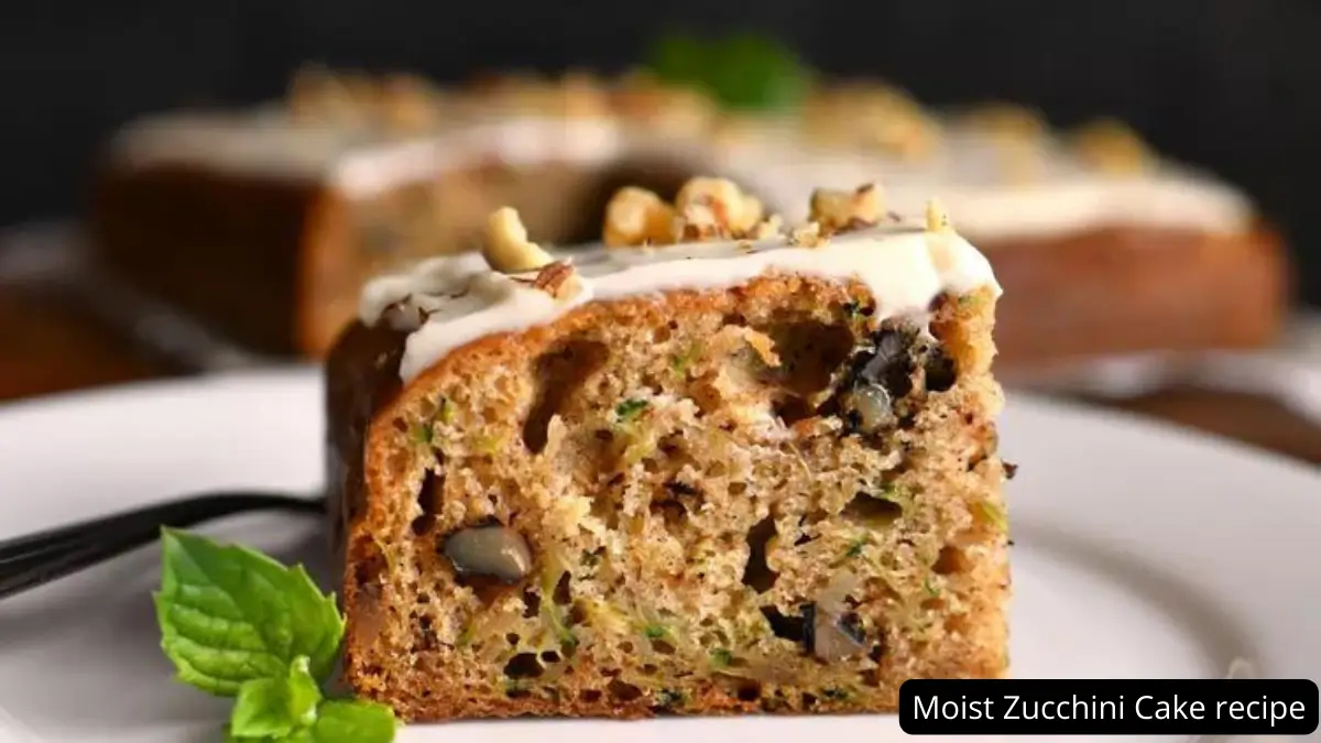 A slice of moist zucchini cake topped with creamy frosting and chopped nuts, served on a white plate with a sprig of fresh mint.