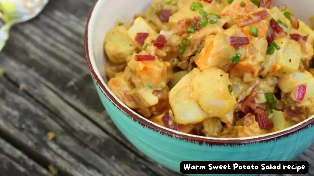 A bowl of warm sweet potato salad with bacon bits and chives on a wooden table.