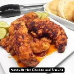 A plate of Nashville Hot Chicken served with golden brown biscuits and pickle slices.