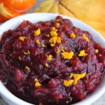 A bowl of crockpot cranberry sauce with apple slices, fresh cranberries, orange zest, and crystallized ginger.